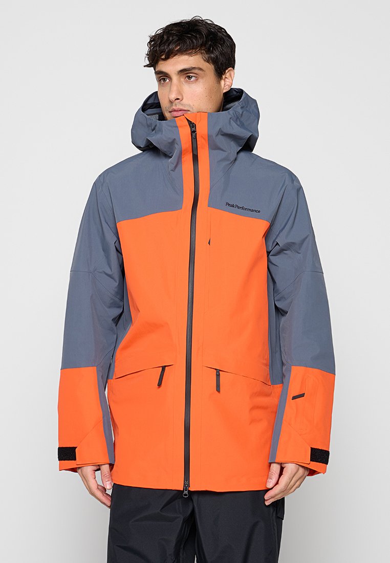 Mann trägt eine zweifarbige orange-graue Kapuzenjacke mit schwarzem Reißverschluss und Taschen, steht vor einem schlichten Hintergrund.