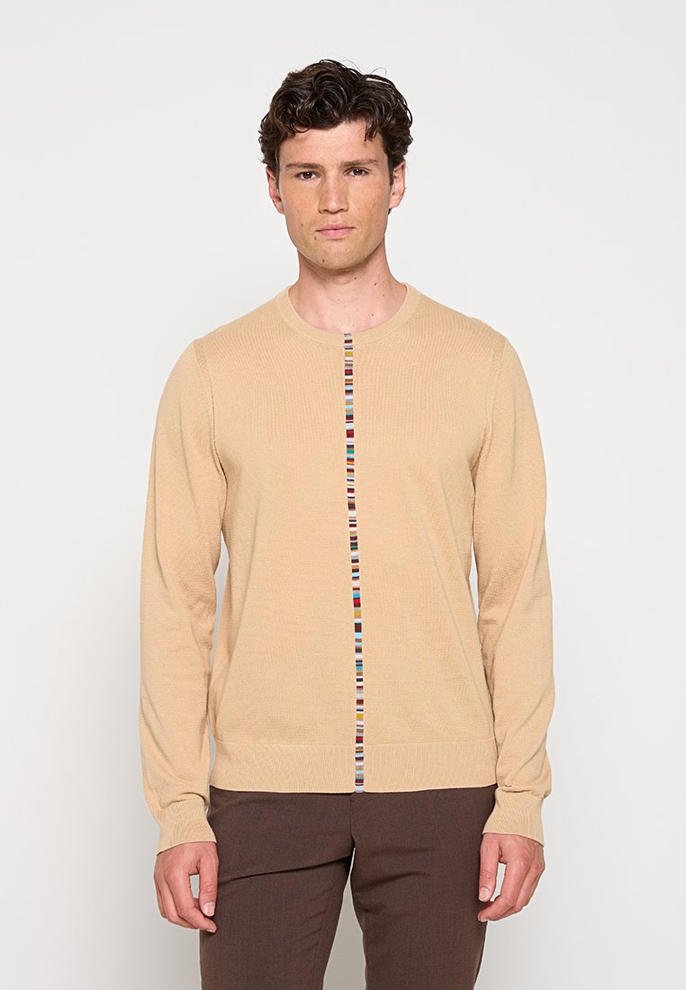 Mann mit beigefarbenem Langarm-Pullover mit vertikalem, mehrfarbigem Streifendetail und brauner Hose steht vor einem schlichten, hellen Hintergrund.