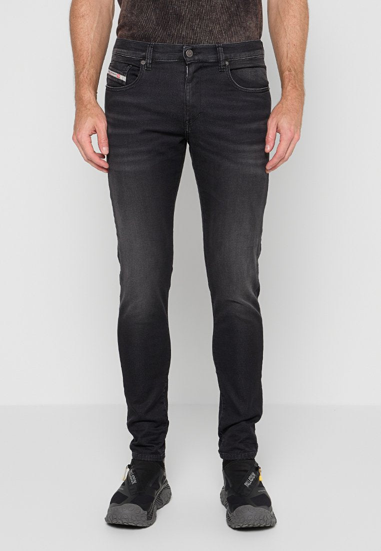 Schlanke schwarze Jeans, getragen von einer Person, die aufrecht steht, kombiniert mit schwarzen Sportschuhen, auf denen der Markenname sichtbar ist.