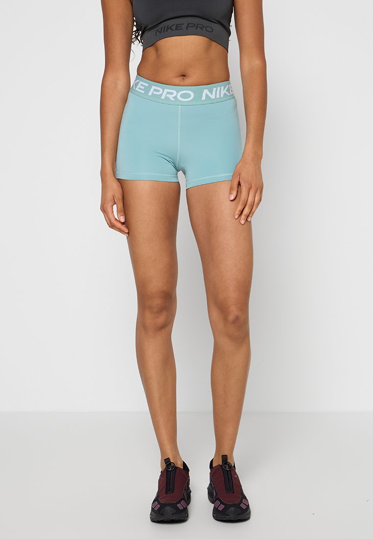 Frau trägt hellblaue Nike Pro Sportshorts und schwarzen bauchfreien Top und steht vor einem einfarbigen weißen Hintergrund.