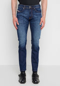 Mann trägt dunkelblaue Slim-Fit-Jeans mit ausgebleichten Details, schwarze Leder-Monk-Strap-Schuhe und ein schwarzes T-Shirt, steht vor einem schlichten Hintergrund.