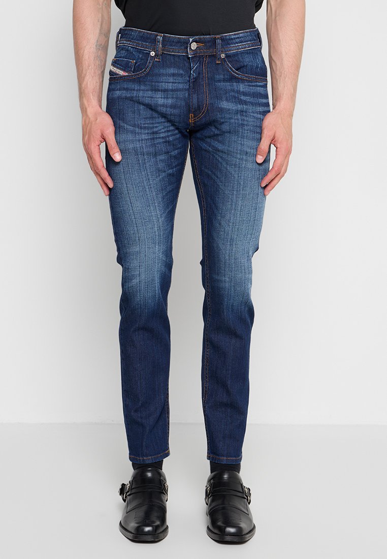 Mann trägt dunkelblaue Slim-Fit-Jeans mit ausgebleichten Details, schwarze Leder-Monk-Strap-Schuhe und ein schwarzes T-Shirt, steht vor einem schlichten Hintergrund.
