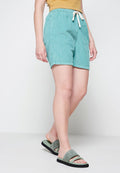 Türkise corduroy Shorts mit einem Kordelzugbund, strukturierten Oberfläche und lockerer Passform, kombiniert mit grünen Slide-Sandalen vor einem neutralen Hintergrund.