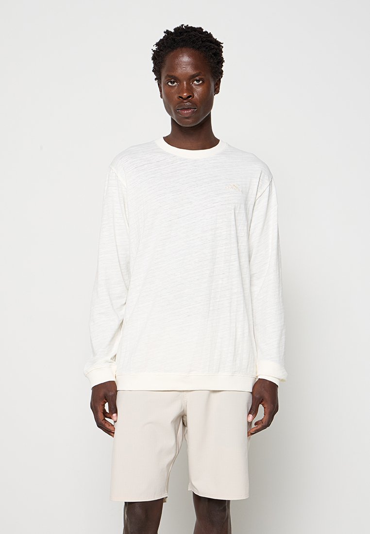 Mann mit kurzen lockigen Haaren, der ein weißes Langarmhemd und beige Shorts trägt, steht vor einem schlichten weißen Hintergrund.
