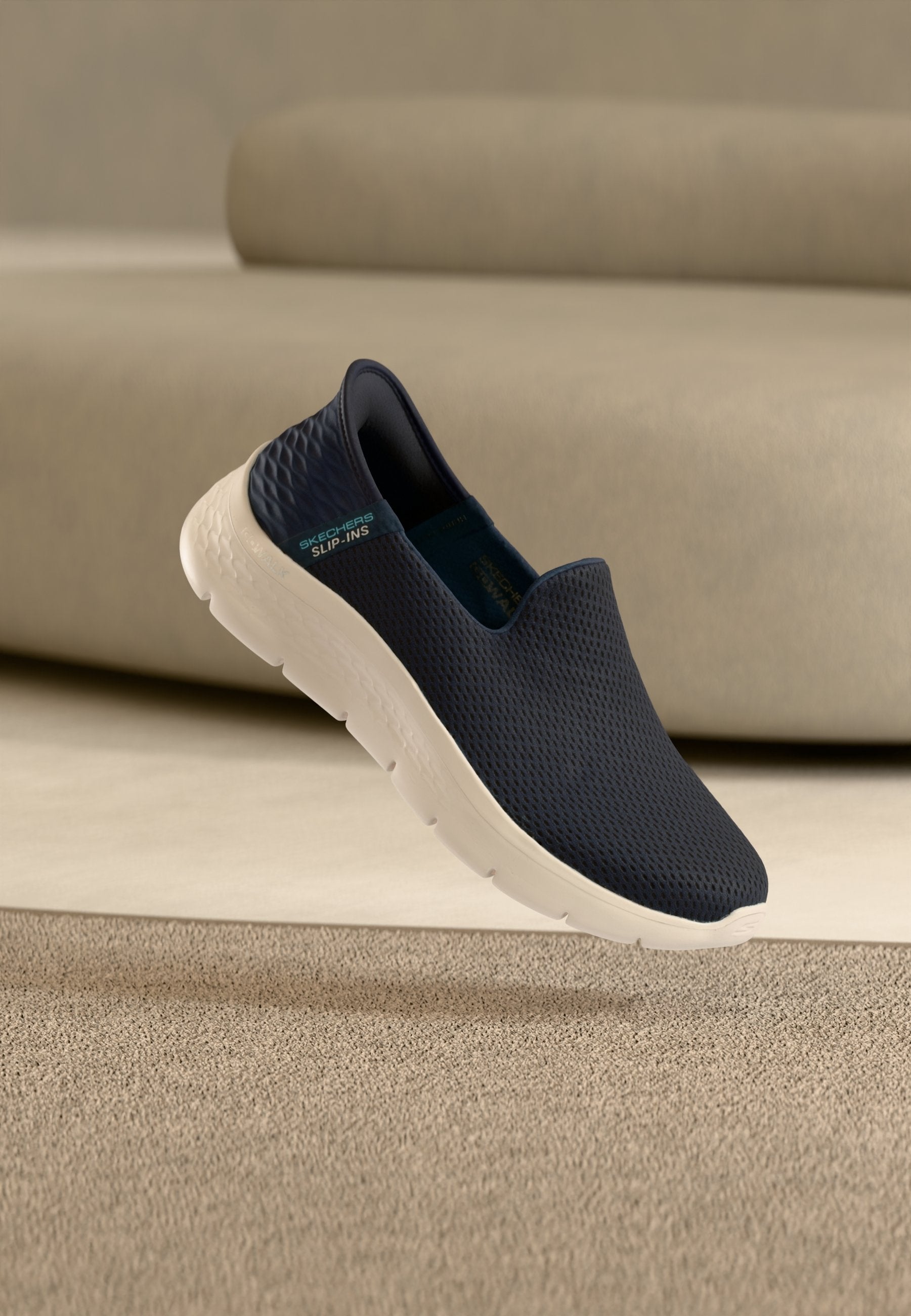 Marineblauer Skechers Slip-On-Sneaker mit weißer Sohle und strukturiertem Obermaterial, schwebt über beigefarbenem Teppich und vor beigefarbenem Sofa.