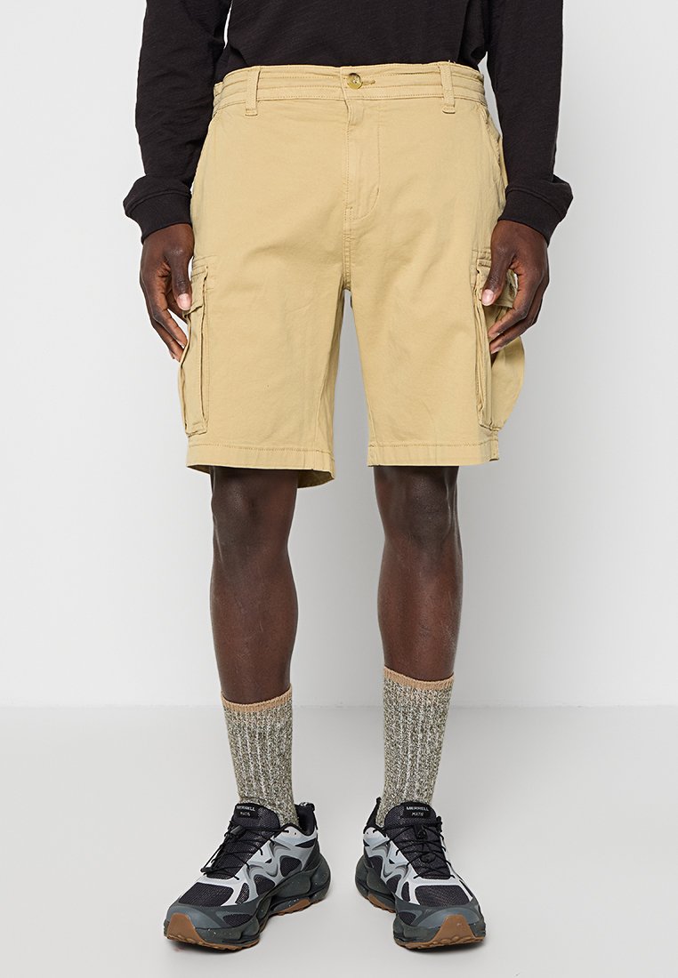 Ein Mann trägt beige Cargo-Shorts, ein schwarzes Langarmshirt, graue gemusterte Socken und schwarz-graue Sportschuhe und steht vor einem schlichten Hintergrund.