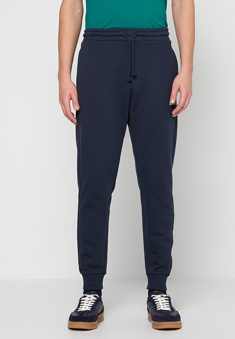 Marineblaue Jogginghosen mit einer gerippten Textur, elastischem Bund und Kordelzug. Verfügen über zulaufende Beine und Seitentaschen. Kombiniert mit dunklen Sneakern.