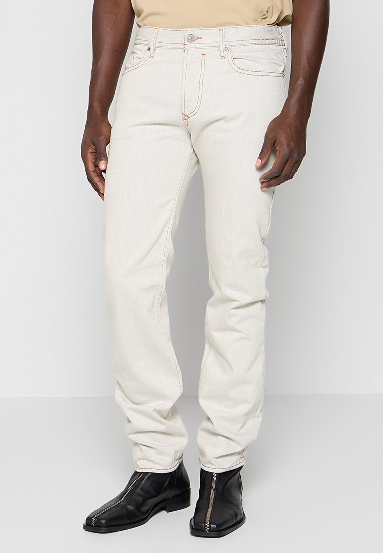 Hellbeige Denim-Jeans mit geradem Bein, sichtbaren Nähten, Fünf-Taschen-Design und einer dezenten Textur. Kombiniert mit schwarzen Stiefeletten.