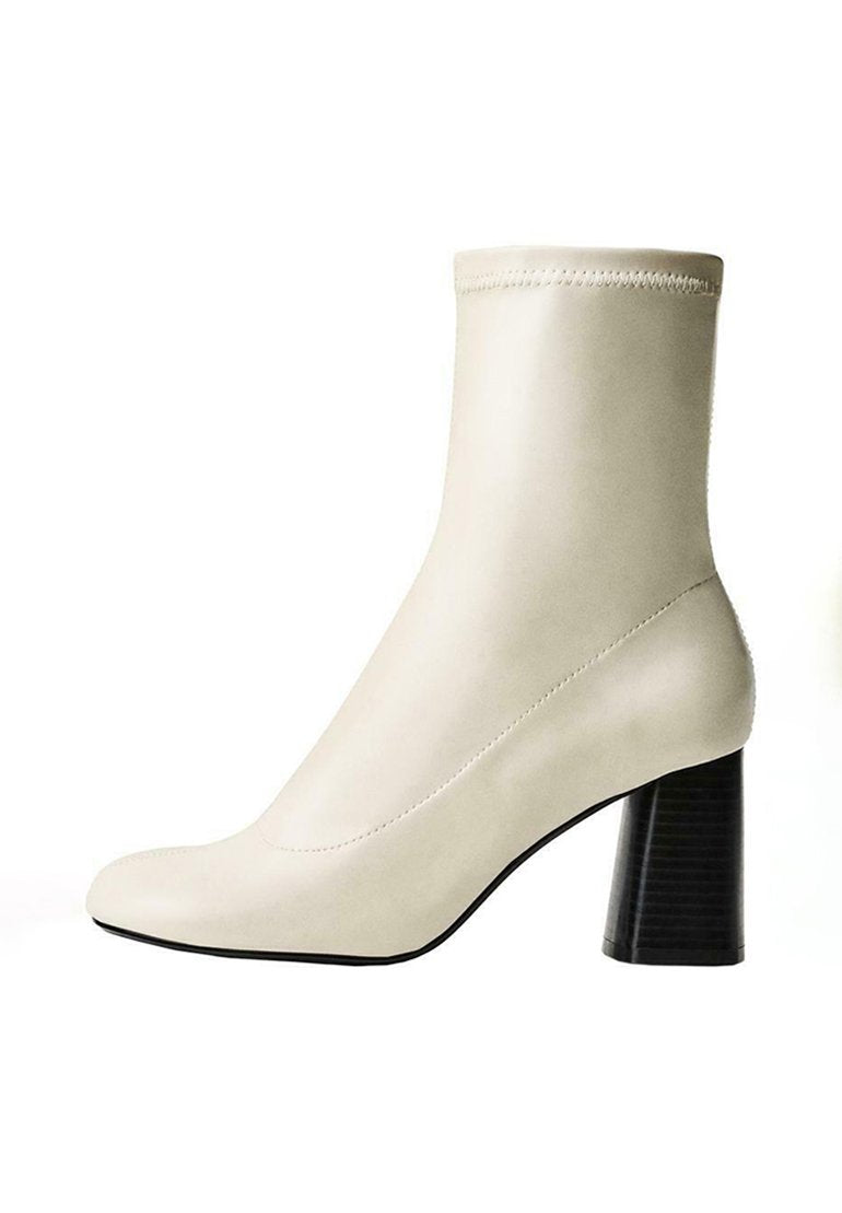Cremefarbene Knöchelstiefel aus glattem Material mit einer spitzen Zehe und einem strukturierten schwarzen Blockabsatz. Nahtdetails entlang der Seiten.