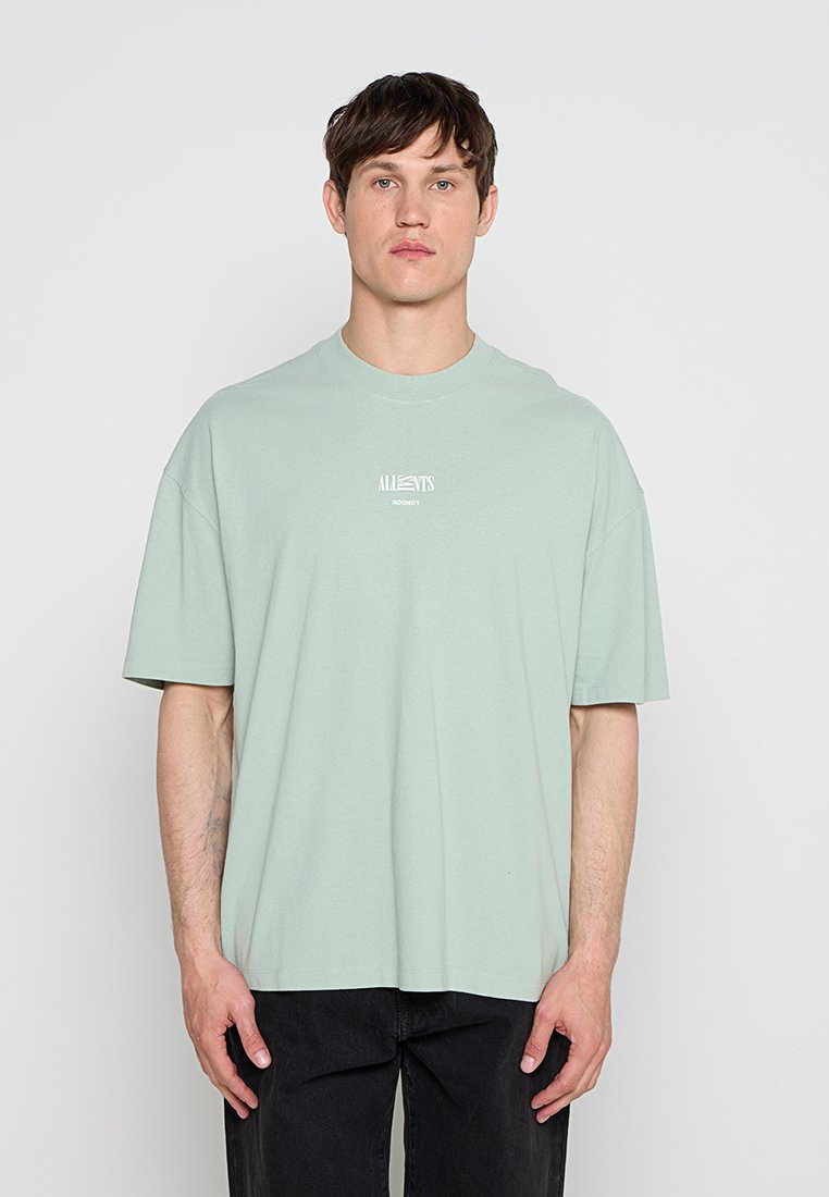 Mann trägt ein hellgrünes oversized T-Shirt mit weißem Logo-Text, steht vor einem schlichten weißen Hintergrund, Hände entspannt seitlich.