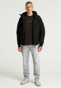 Schwarze gepolsterte Jacke mit hohem Kragen, Reißverschluss und Logodetail. Kombiniert mit einem dunklen T-Shirt und hellen Jeans, ergänzt durch beige Sneakers.