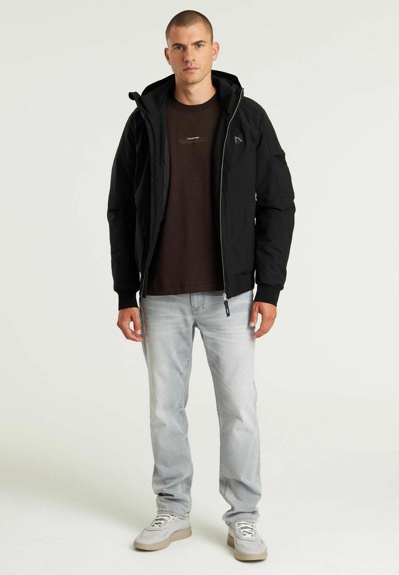 Schwarze gepolsterte Jacke mit hohem Kragen, Reißverschluss und Logodetail. Kombiniert mit einem dunklen T-Shirt und hellen Jeans, ergänzt durch beige Sneakers.