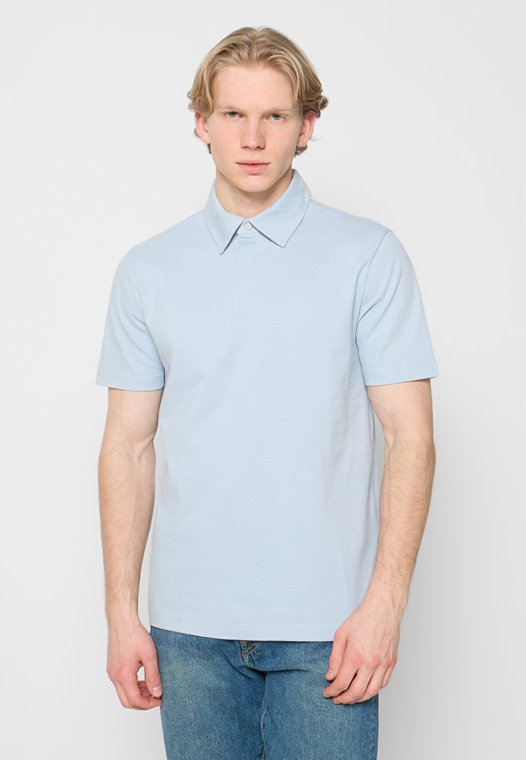 Junger Mann mit blondem Haar, der ein hellblaues kurzärmeliges Poloshirt und blaue Jeans vor einem einfarbigen weißen Hintergrund trägt.