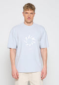 Blonder Mann trägt hellblaues oversized T-Shirt mit weißem kreisförmigem Text und beiger Hose, steht vor einem weißen Hintergrund.
