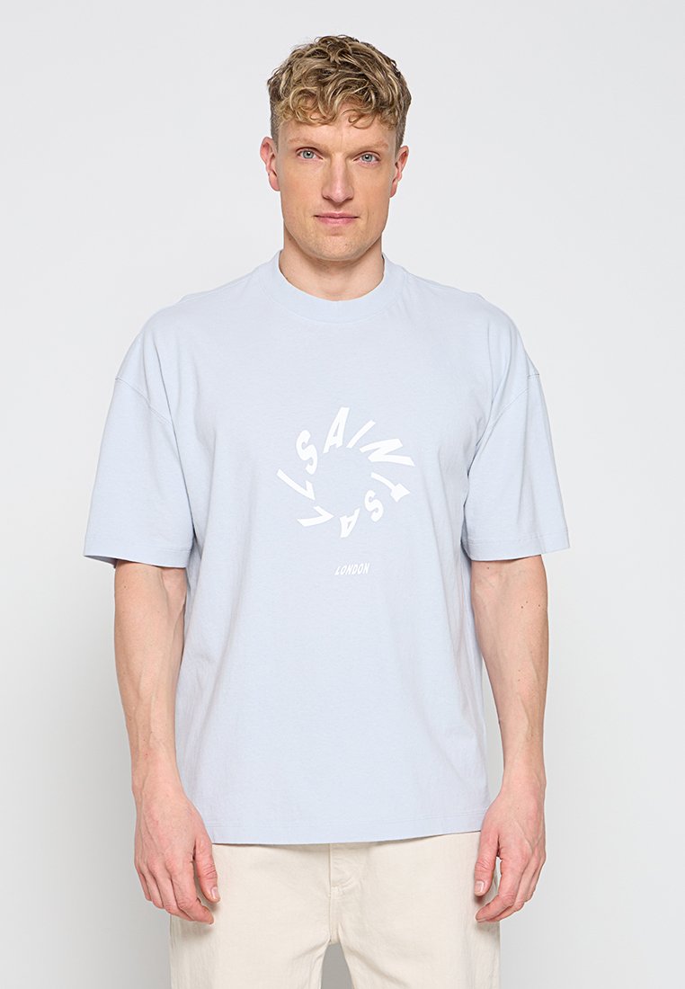 Blonder Mann trägt hellblaues oversized T-Shirt mit weißem kreisförmigem Text und beiger Hose, steht vor einem weißen Hintergrund.