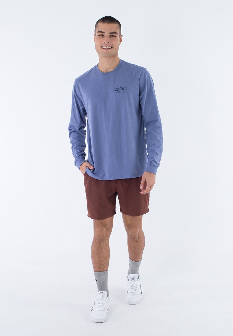 Langärmliges blaues Hemd mit kleinem Logo, kombiniert mit braunen Shorts. Weiße Sneakers und graue Socken runden das Outfit ab, präsentiert auf einem schlichten Hintergrund.
