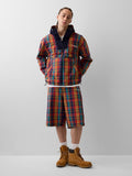 Person steht, trägt eine mehrfarbige karierte Jacke und passende Shorts mit beigen Arbeitsschuhen und weißen Socken vor einem schlichten grauen Hintergrund.