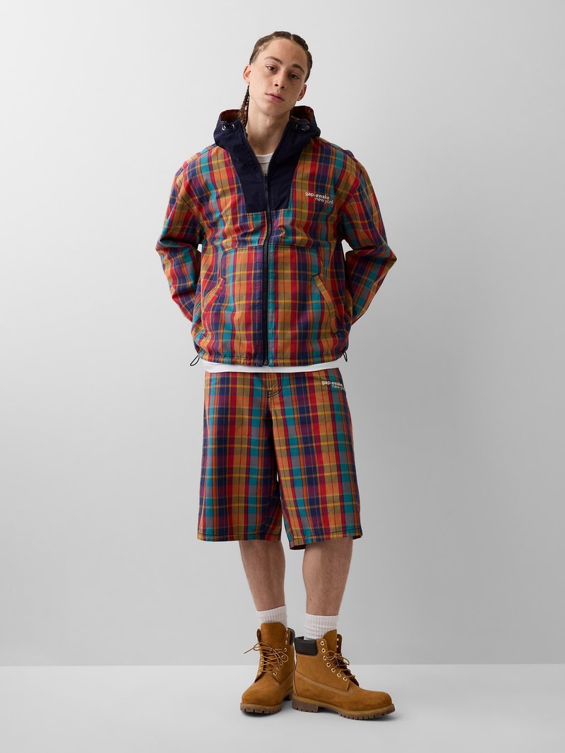 Person steht, trägt eine mehrfarbige karierte Jacke und passende Shorts mit beigen Arbeitsschuhen und weißen Socken vor einem schlichten grauen Hintergrund.