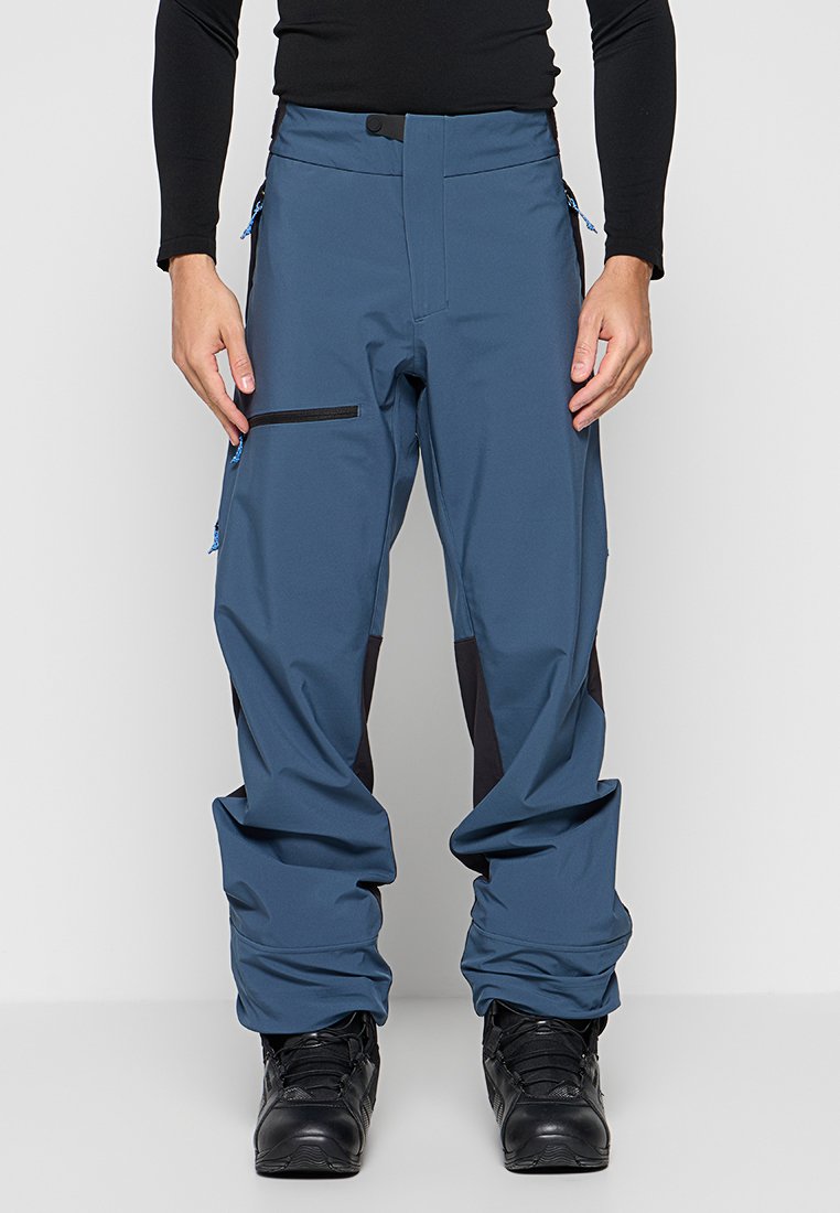 Mann trägt blaue Cargohosen mit schwarzen Kniepolstern und schwarzen Stiefeln, steht vor einem schlichten hellen Hintergrund.