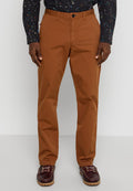 Mann in rostfarbenen Chinos mit einem schwarzen gemusterten Hemd und dunkelbrown Leder-Schuhen, stehend vor einem einfarbigen Hintergrund.