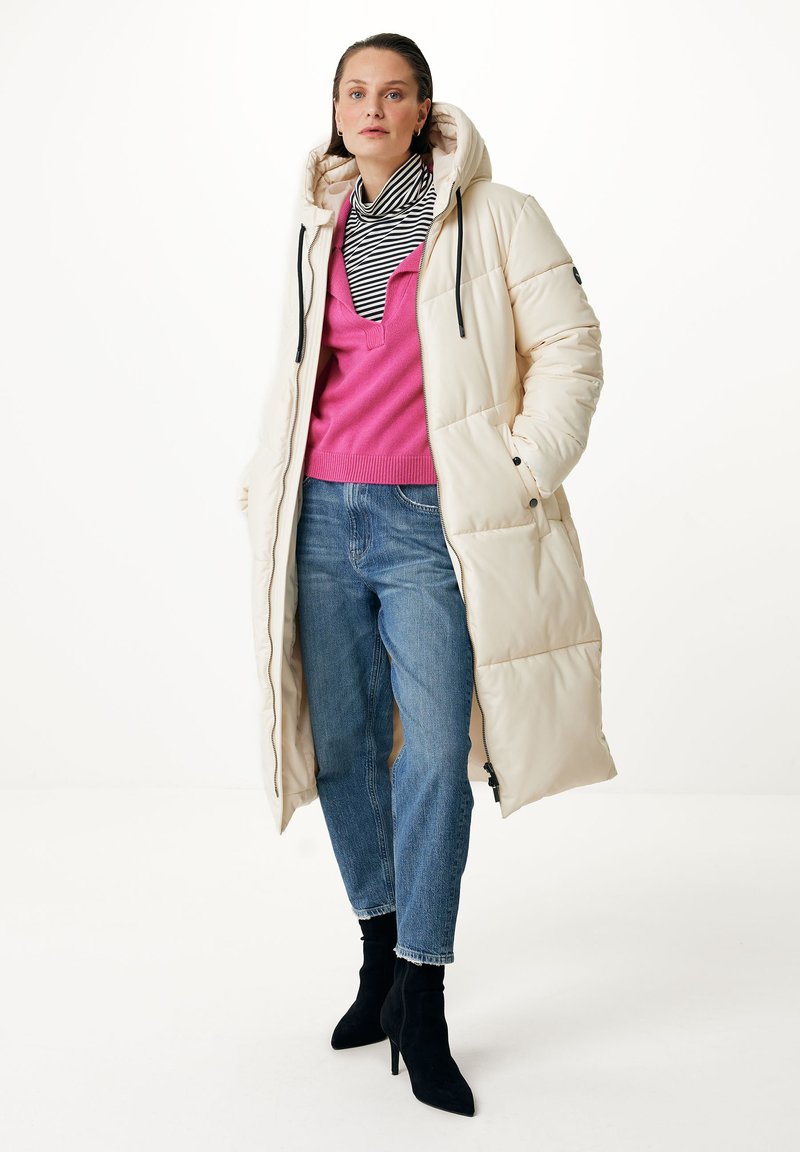 Cremfarbener Steppmantel mit Kapuze, ausgestattet mit schwarzen Zugbändern, kombiniert mit einem pinken Pullover und blauen Jeans. Schwarze Knöchelstiefel runden das Outfit ab.
