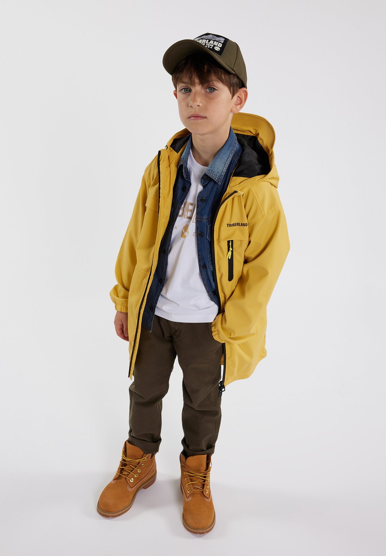 Gelbe wasserdichte Jacke mit schwarzer Innenausstattung, über einem Denim-Hemd getragen. Olivgrüne Hose und braune Stiefel mit gelben Schnürsenkeln vervollständigen das Outfit.