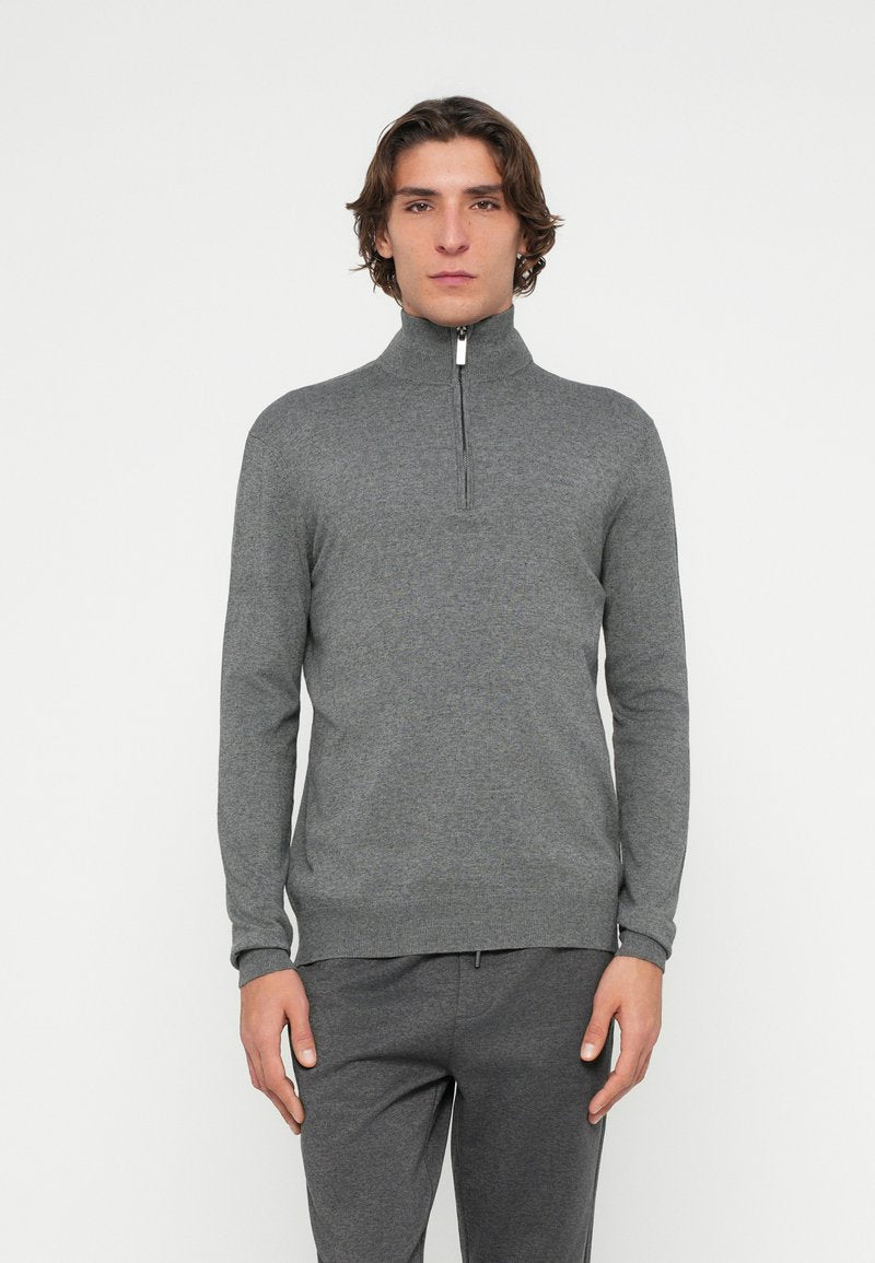 Grau melierter Pullover mit Reißverschluss bis zur Brust, langen Ärmeln, strukturiertem Strickstoff und weißem Reißverschlussdetail am Kragen. Kombiniert mit passenden grauen Hosen.