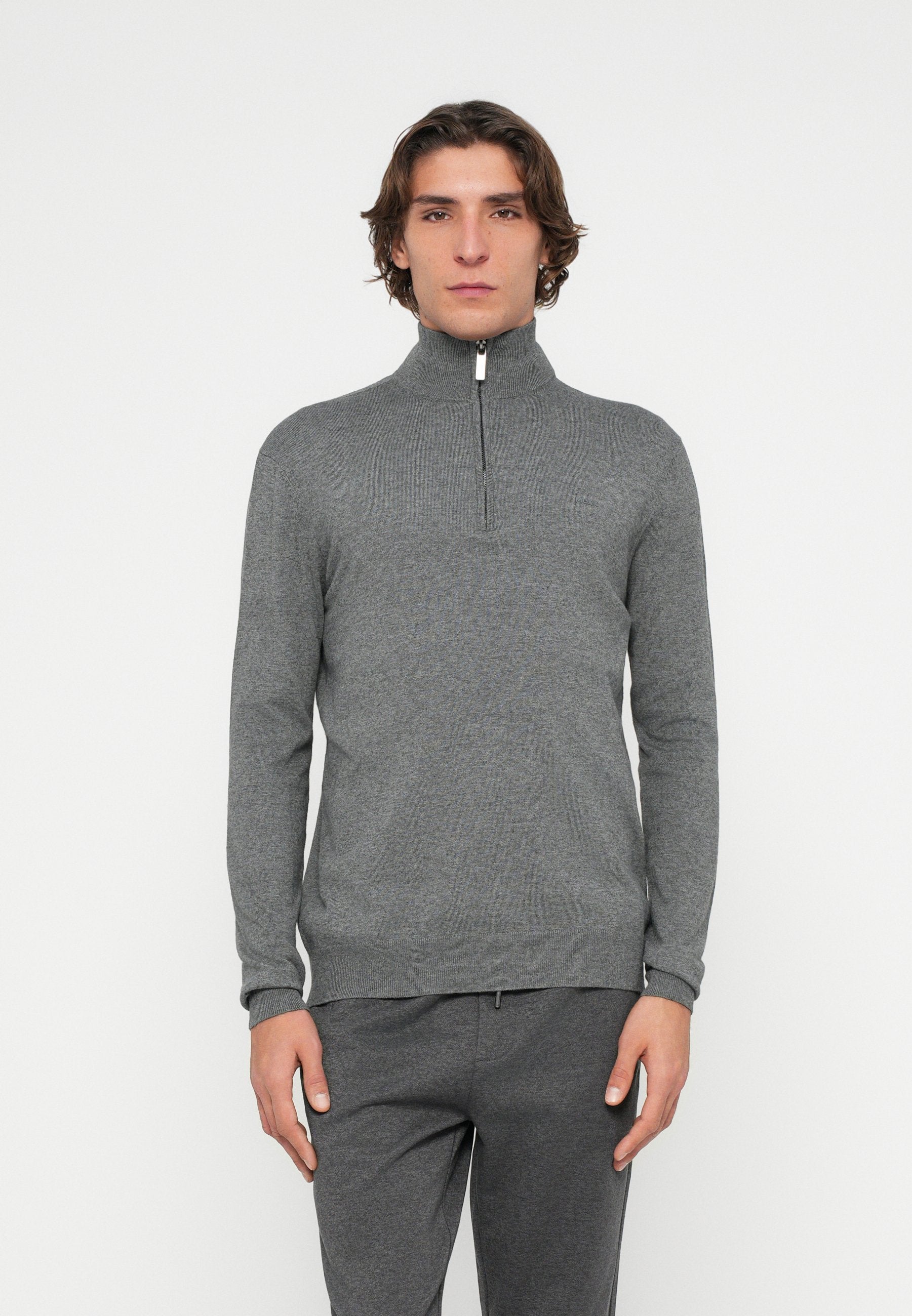 Grau melierter Pullover mit Reißverschluss bis zur Brust, langen Ärmeln, strukturiertem Strickstoff und weißem Reißverschlussdetail am Kragen. Kombiniert mit passenden grauen Hosen.
