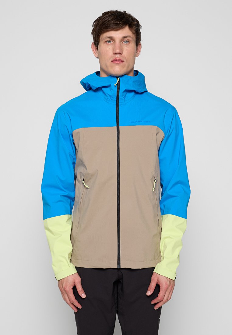 Mann trägt eine blau-beige-hellgelbe Kapuzenjacke mit Reißverschluss und vorderen Reißverschlusstaschen, steht vor einem schlichten grauen Hintergrund.