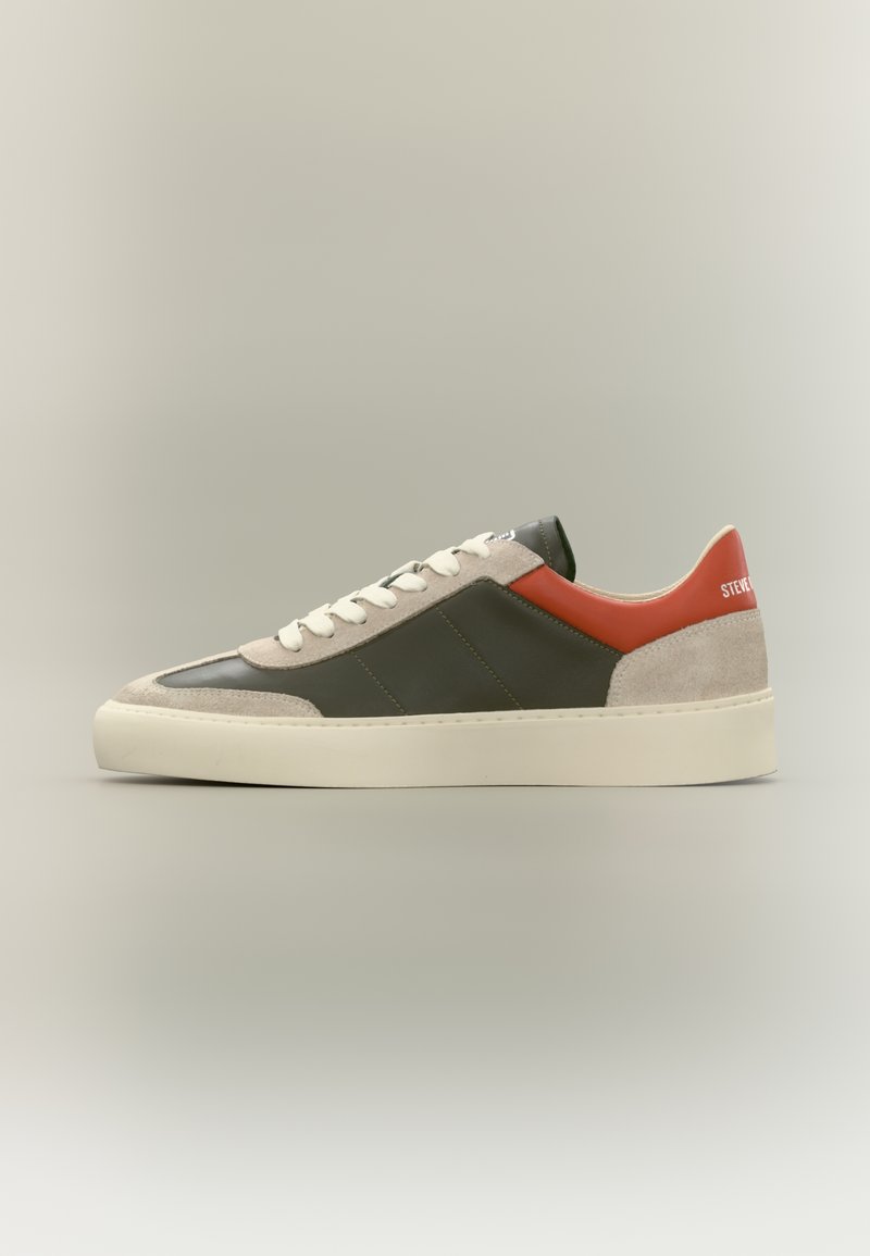 Flacher Sneaker aus dunkelgrünem Leder mit beigen Wildlederakzenten, weißen Schnürsenkeln, rotem Fersen-Detail und cremefarbener Sohle auf schlichten Hintergrund.
