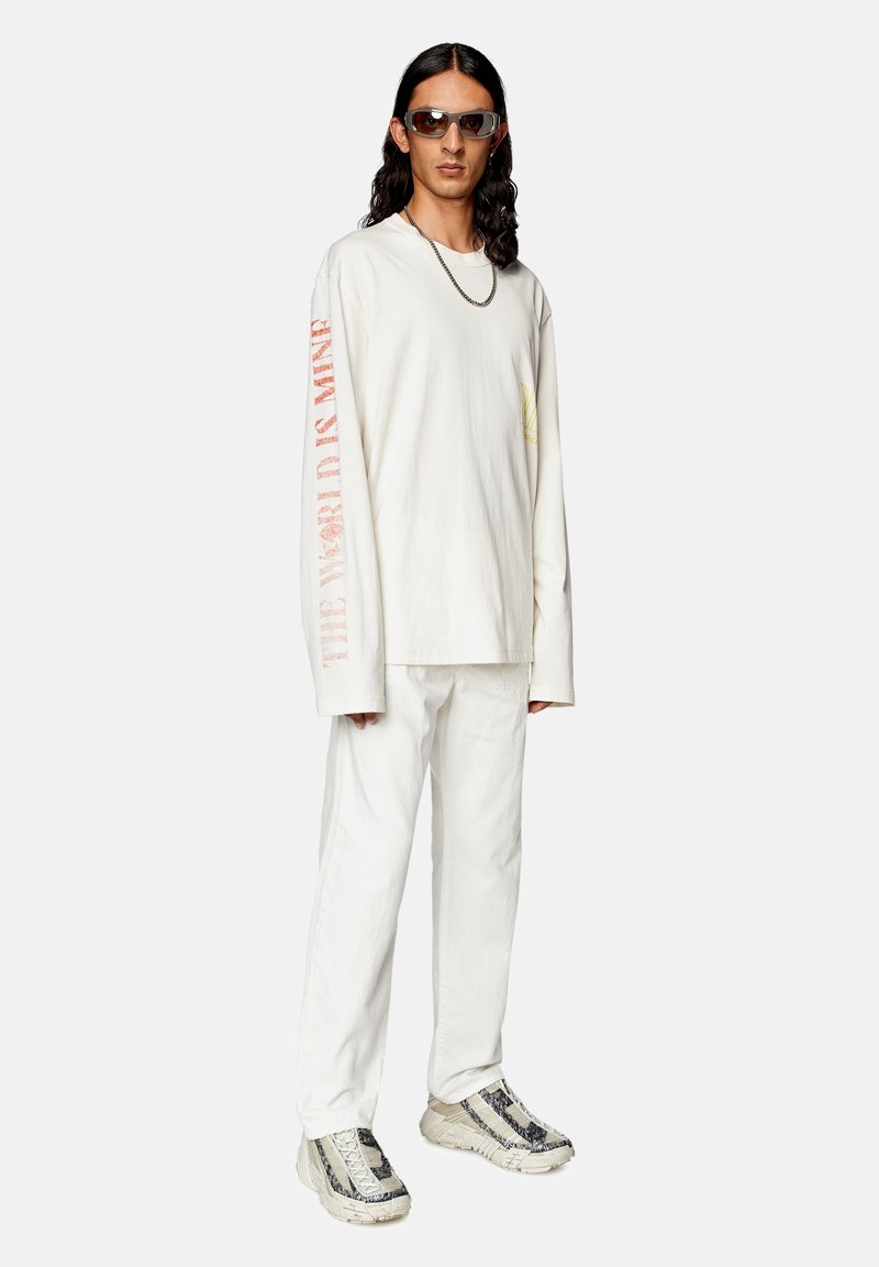 Junger Mann mit langen Haaren, der ein übergroßes weißes Langarmshirt mit rotem Schriftzug an den Ärmeln, weiße Hosen, gemusterte Sneakers, eine Kettenhalskette und Sonnenbrillen trägt.