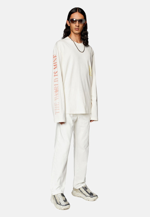 Junger Mann mit langen Haaren, der ein übergroßes weißes Langarmshirt mit rotem Schriftzug an den Ärmeln, weiße Hosen, gemusterte Sneakers, eine Kettenhalskette und Sonnenbrillen trägt.