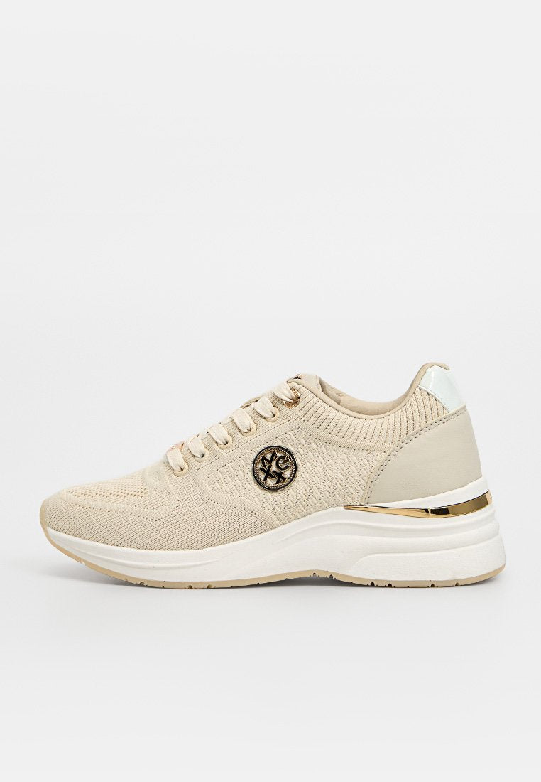 Beige gestrickter Sneaker mit weißer Sohle, goldfarbenem Akzent an der Ferse, Schnürung vorne und rundem Logo-Emblem an der Seite.