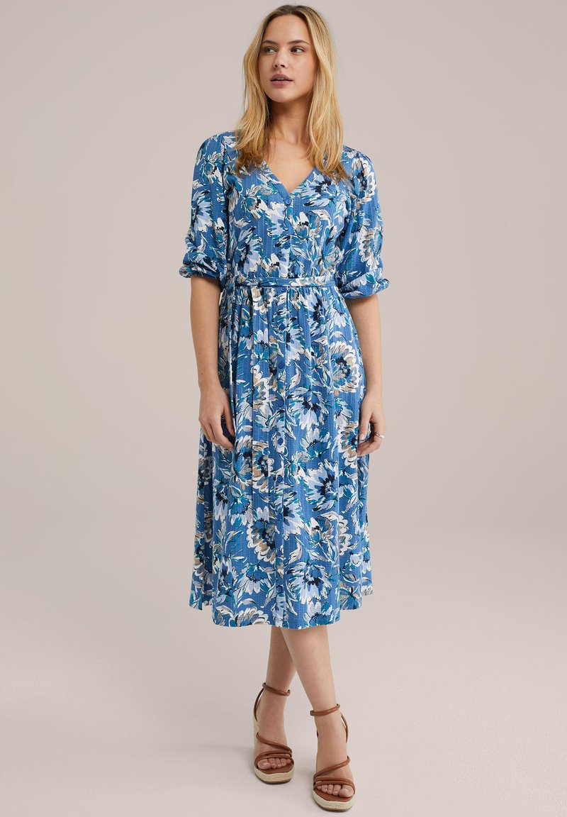 Blaues Blumenkleid in Midilänge mit kurzen Puffärmeln, V-Ausschnitt und Bindeband in der Taille. Das Kleid besteht aus einem gemusterten Stoff in Blau- und Weißtönen.
