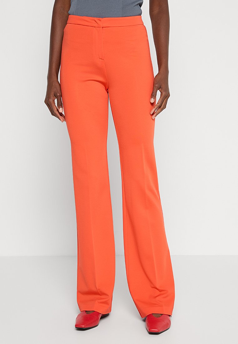 Leuchtend orangefarbene Hosen mit glatter Textur, ausgestelltem Design, mittelhohem Bund und geradem Schnitt. Kombiniert mit roten Schuhen.