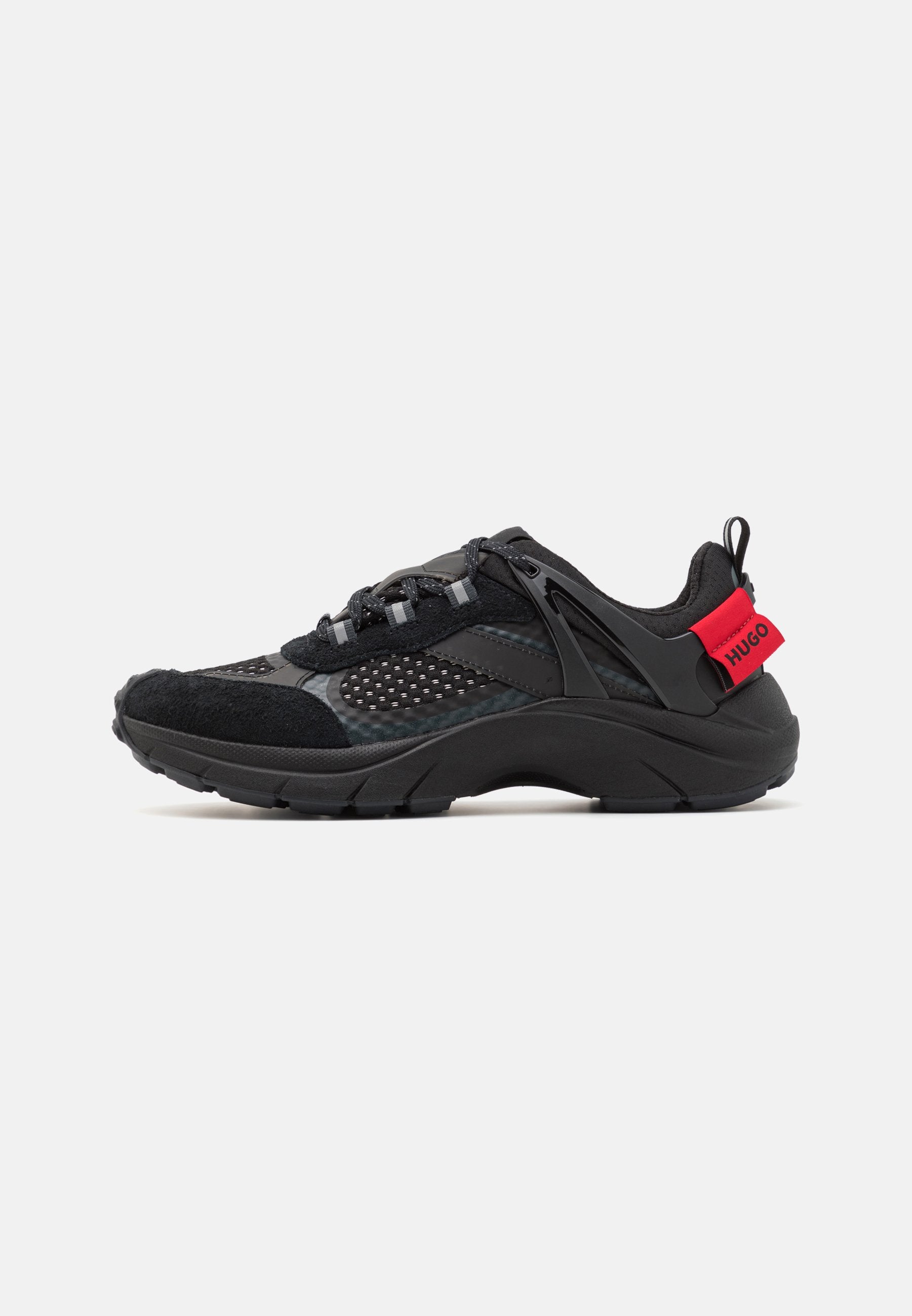 Schwarzer Sportschuh aus Mesh und Wildleder, mit einer dicken Sohle, geschwungenem Design, reflektierenden Akzenten und einer roten Markenlasche am Fersenbereich.
