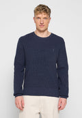 Blonder Mann mit einem dunkelblauen strukturierten Strickpullover und hellbeigen Hosen, der vor einem schlichten hellgrauen Hintergrund steht.