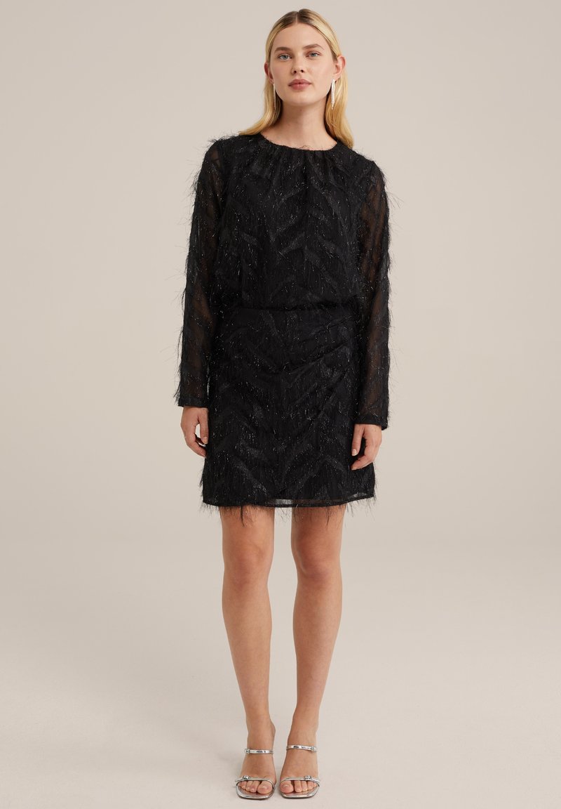 Schwarzes Kleid mit langen transparenten Ärmeln, strukturiert mit einem Zickzack-Muster, mit taillierter Taille und fransigem Saum. Kombiniert mit silbernen Sandalen.