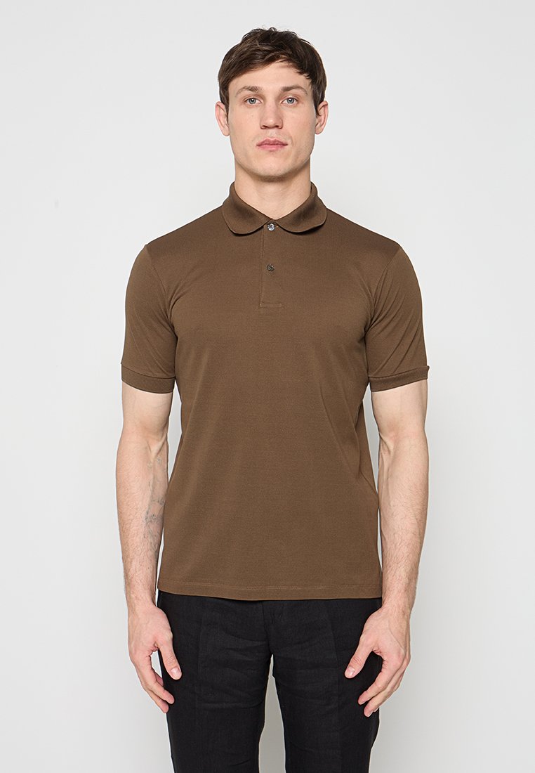 Mann steht frontal, trägt ein braunes kurzärmeliges Poloshirt und eine schwarze Hose vor einem schlichten hellen Hintergrund.