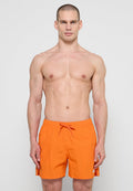 Muskulöser Mann mit kurzen Haaren steht vor einfarbigem Hintergrund und trägt knallorange Badehosen mit Kordelzug in der Taille.