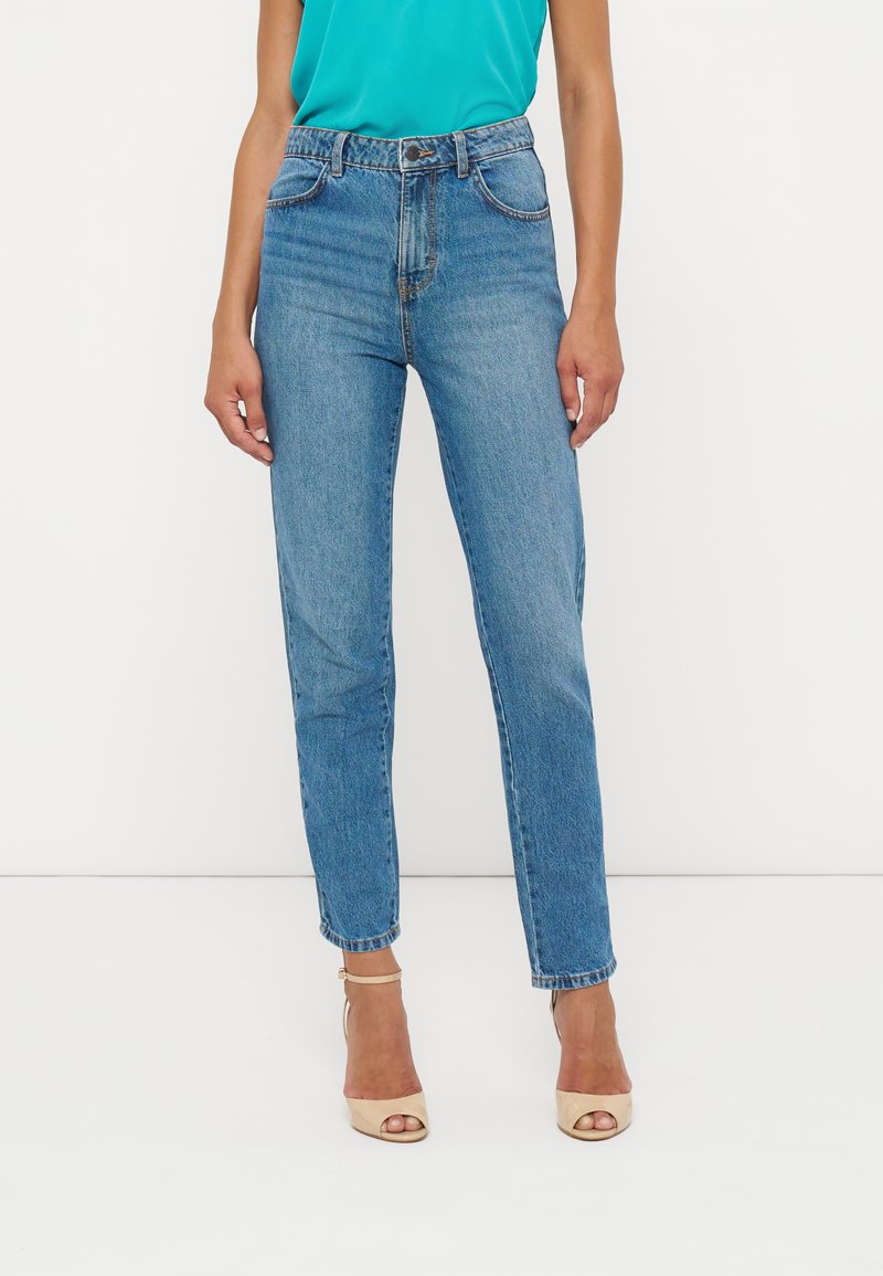 Frau mit hoch taillierten blauen Jeans und beigen offenen High Heels, stehend vor einem schlichten weißen Hintergrund.