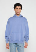 Mann mit lockigem Haar, der einen hellblauen, übergroßen Hoodie und hellblaue Jeans trägt, vor einem schlichten, hellen Hintergrund steht.