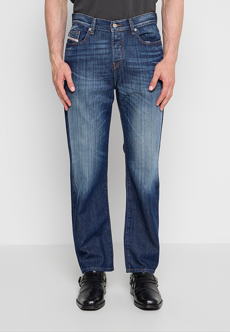 Ein Mann trägt blaue ausgewaschene Straight-Leg-Jeans und schwarze Ledermokassins mit Riemen und steht vor einem schlichten hellen Hintergrund.