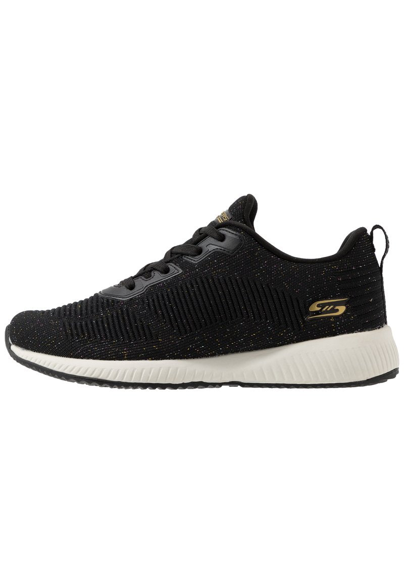 Schwarzer Sportschuh mit einem strukturierten Strick-Obermaterial, das metallische Sprenkel aufweist. Dicke weiße Zwischensohle und schwarze Außensohle. Goldenes Logo-Detail an der Seite.