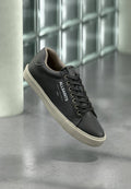 Schwarzer Ledersneaker von AllSaints mit weißer Sohle, schwarzen Schnürsenkeln und Logo an der Seite, schwebt über einem grau reflektierenden Boden.