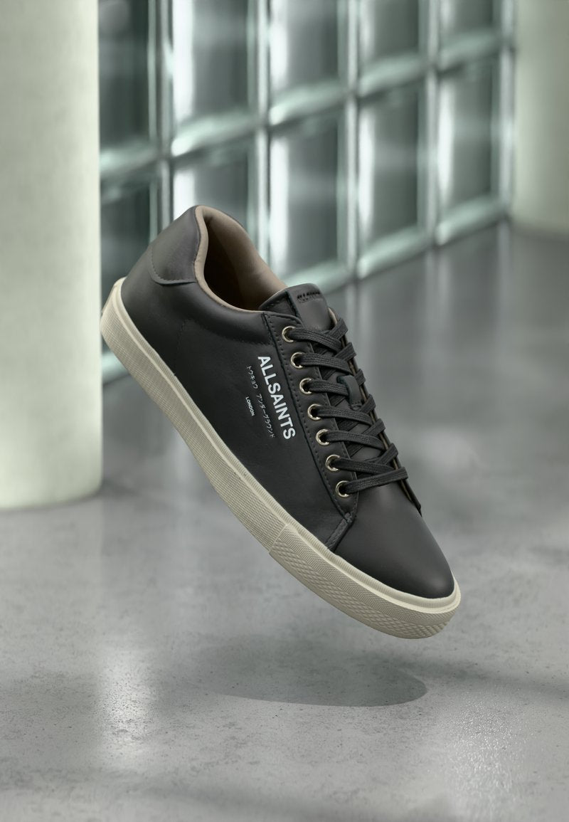 Schwarzer Ledersneaker von AllSaints mit weißer Sohle, schwarzen Schnürsenkeln und Logo an der Seite, schwebt über einem grau reflektierenden Boden.