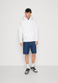 Junger Mann, der einen weißen Hoodie, blaue Jeansshorts, schwarz-weiße Turnschuhe, weiße Socken und eine schwarze Mütze trägt und vor einer schlichten weißen Wand steht.