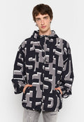 Junger Mann in einer schwarz-grau gemusterten Kapuzenjacke mit großen abstrakten Buchstaben, die Hände in den Taschen, vor einem weißen Hintergrund.