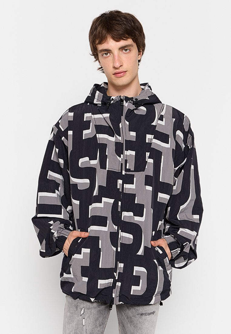 Junger Mann in einer schwarz-grau gemusterten Kapuzenjacke mit großen abstrakten Buchstaben, die Hände in den Taschen, vor einem weißen Hintergrund.