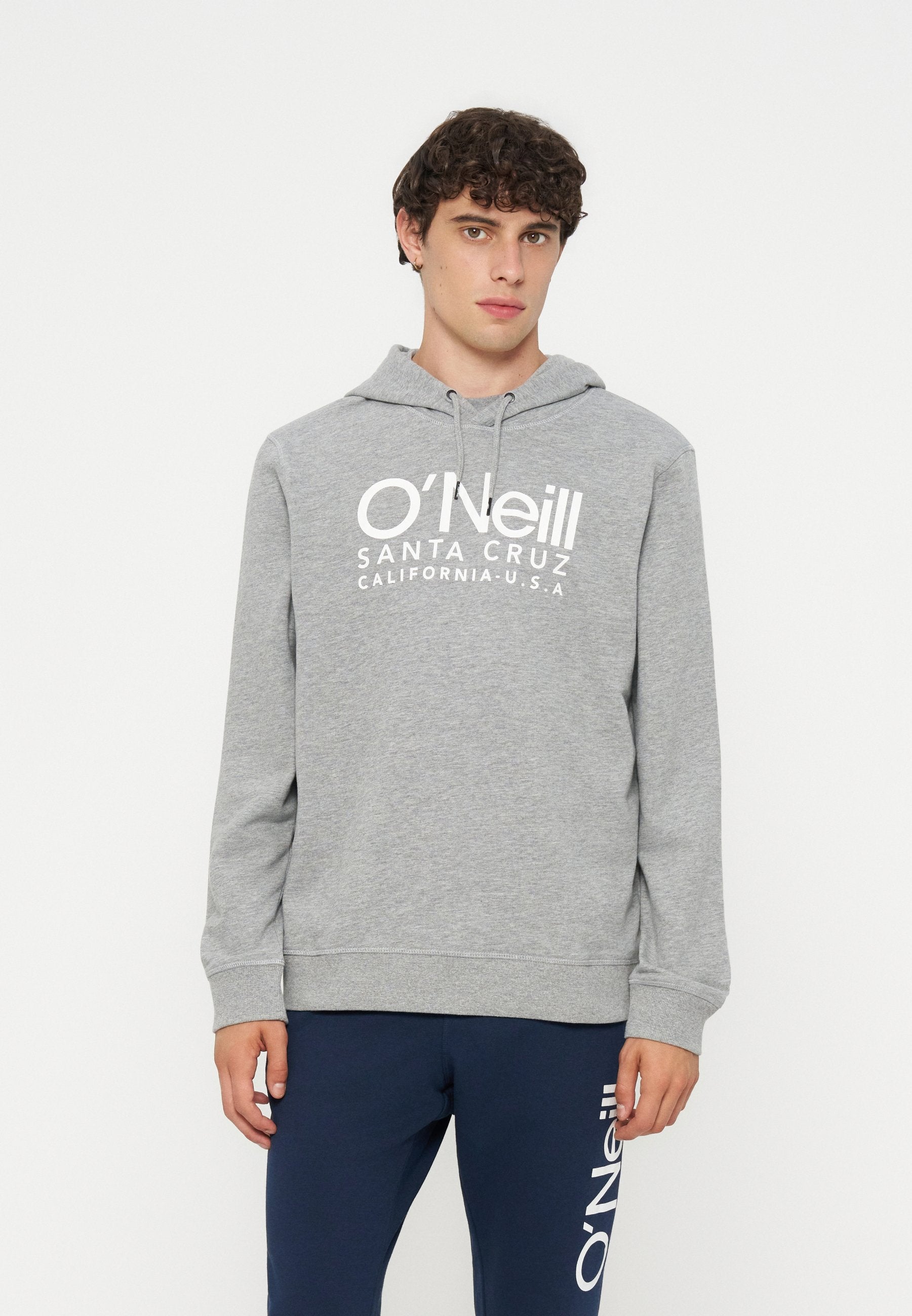 Junger Mann mit lockigem Haar, der einen grauen O’Neill-Hoodie und marineblaue Hose mit O’Neill-Logo auf einem Bein trägt, steht vor einem schlichten Hintergrund.
